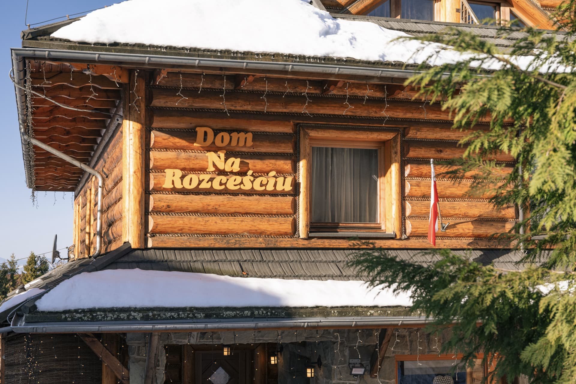 The log facade in winter with snow on the roof, string lights and the carved house sign